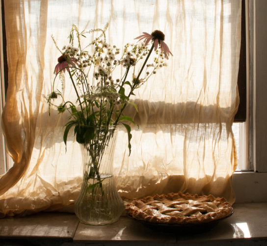 A lattice top cherry pie and vase filled with wildflowers near an open window on the farm.