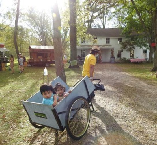 A man pulls a cart with smiling children toward the taco tent at Fermentation Fest. Artist-made roadside culture stands are seen in the distance.